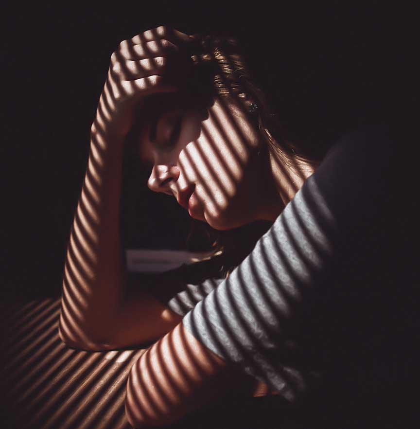 A distressed woman sits alone in a dark room lit through venetian blinds